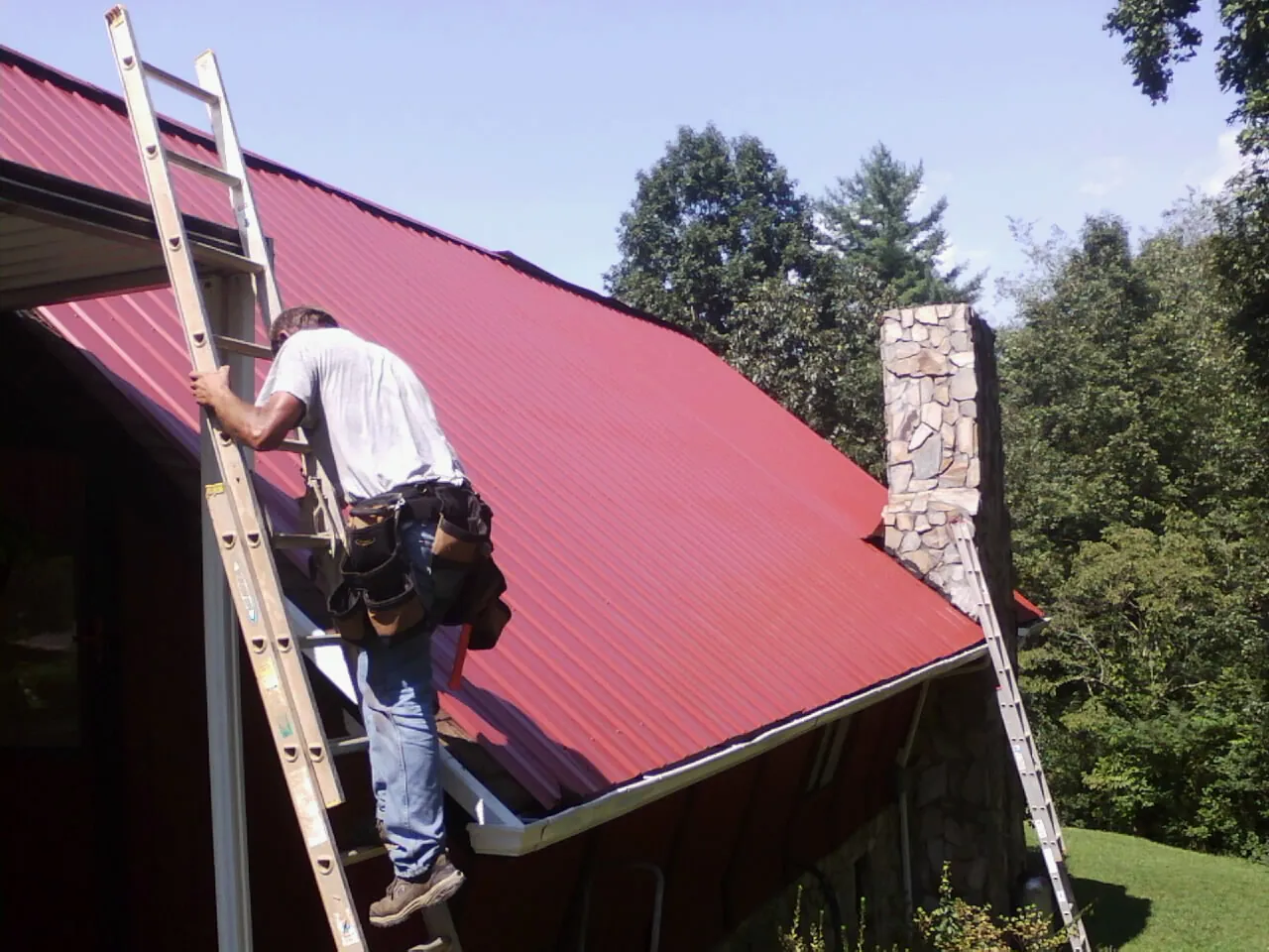 Roofer on a ladder beside a red metal roof and stone chimney — Soffit & Fascia Repair by Roofing Brothers in Casa Blanca